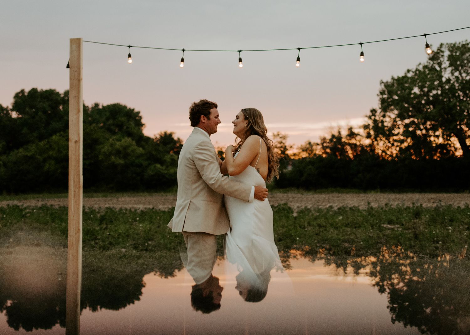 A romantic couple shares an intimate moment at sunset with string lights and a reflective pond in the background.