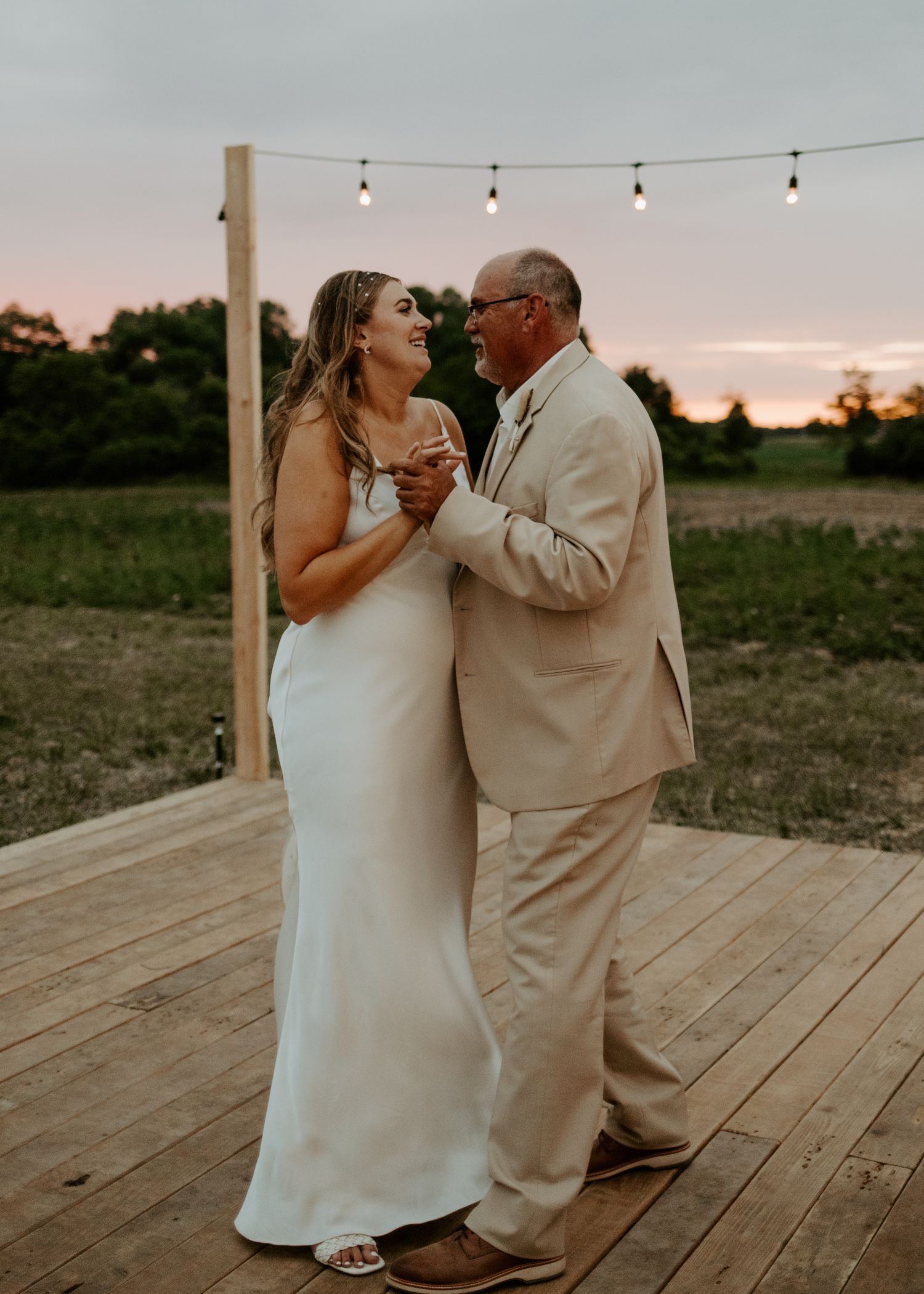Couple shares an intimate dance on a wooden platform under string lights at sunset.