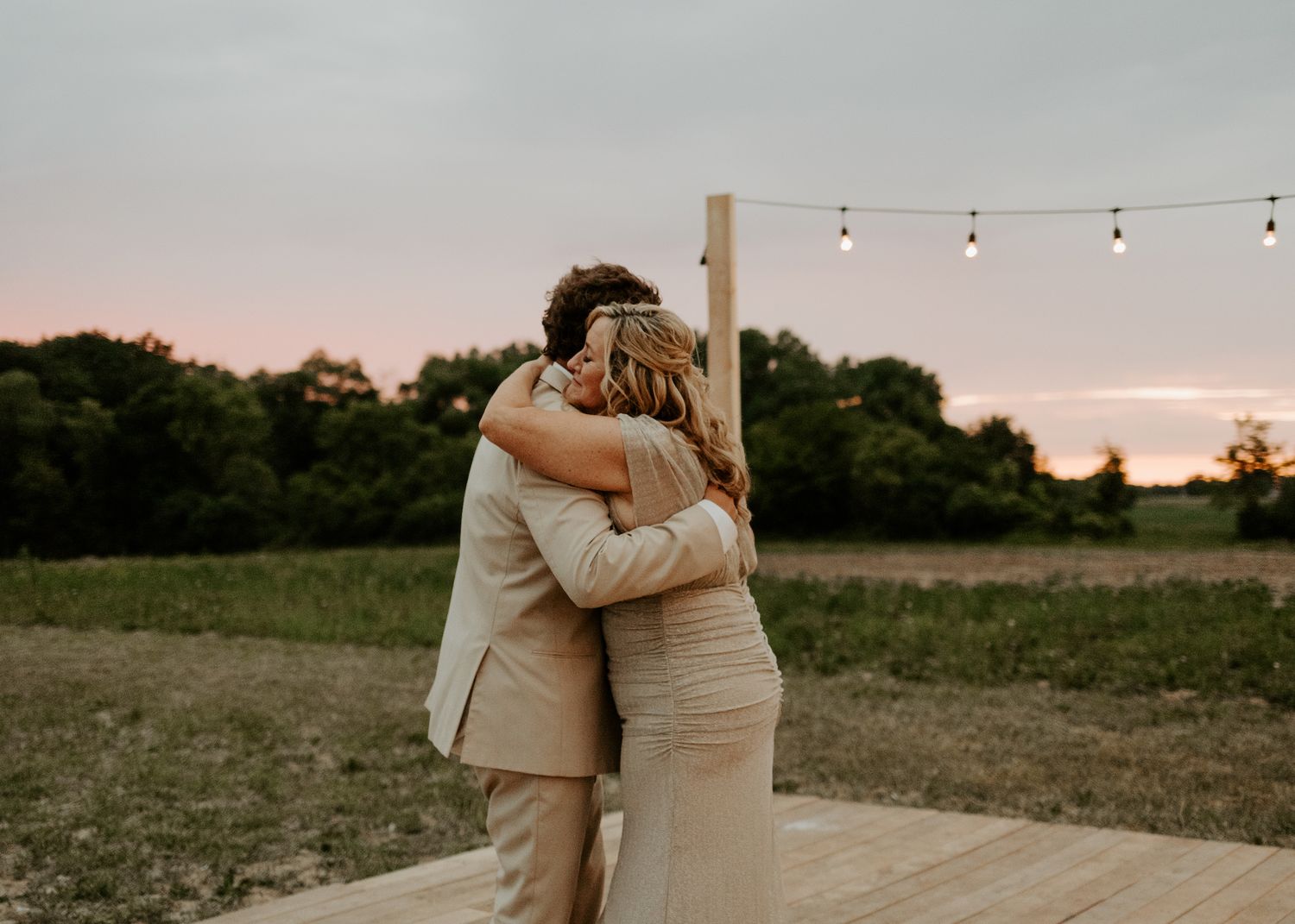 Newlyweds share a tender dance moment on a wooden deck at dusk under string lights.