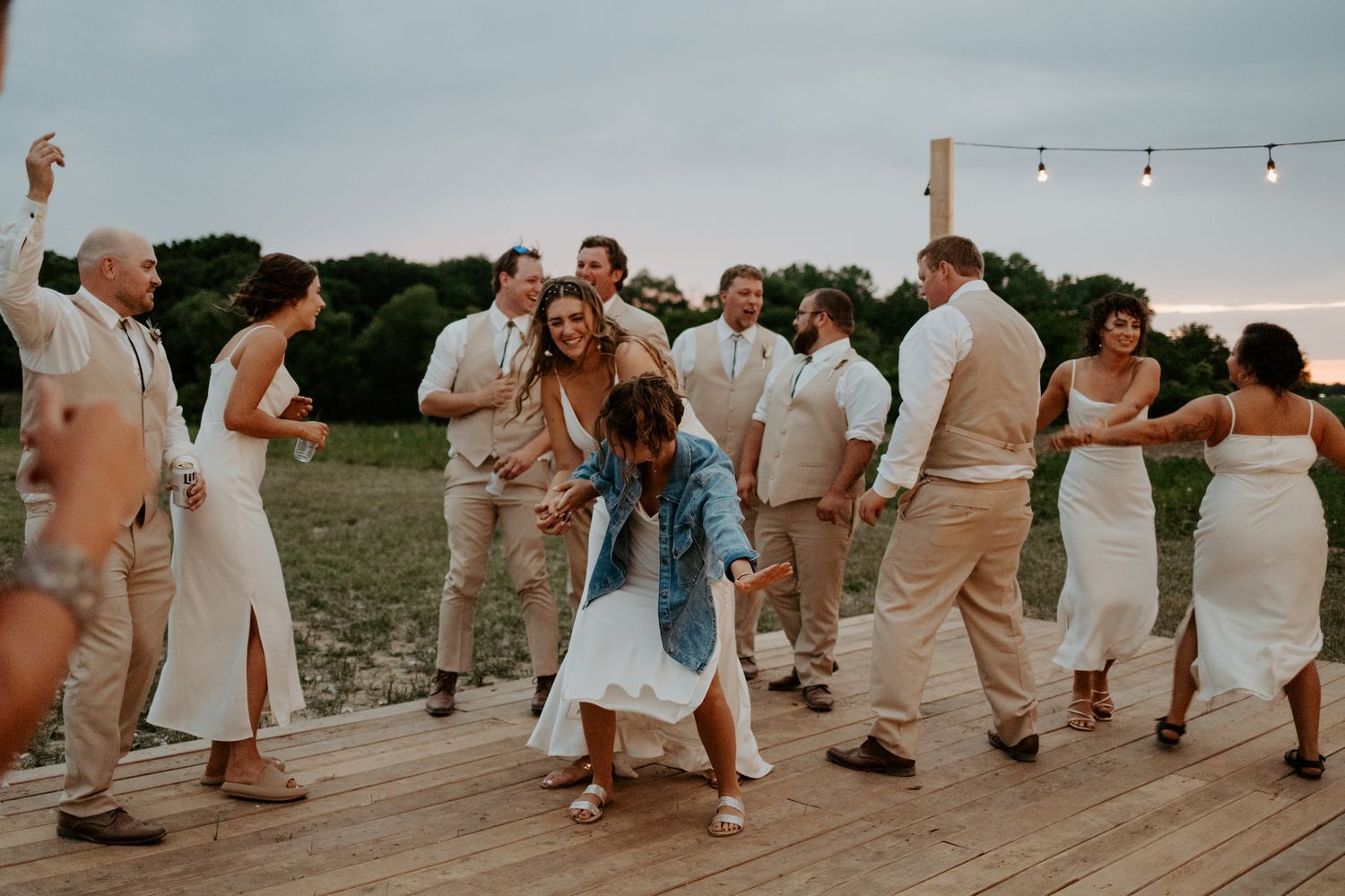 Wedding party dances and celebrates on an outdoor wooden deck under string lights at dusk.
