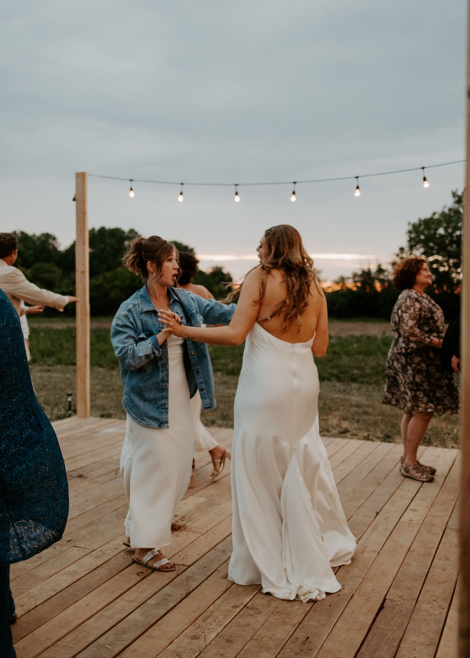Wedding celebration with dancing guests on a wooden deck at sunset under string lights.