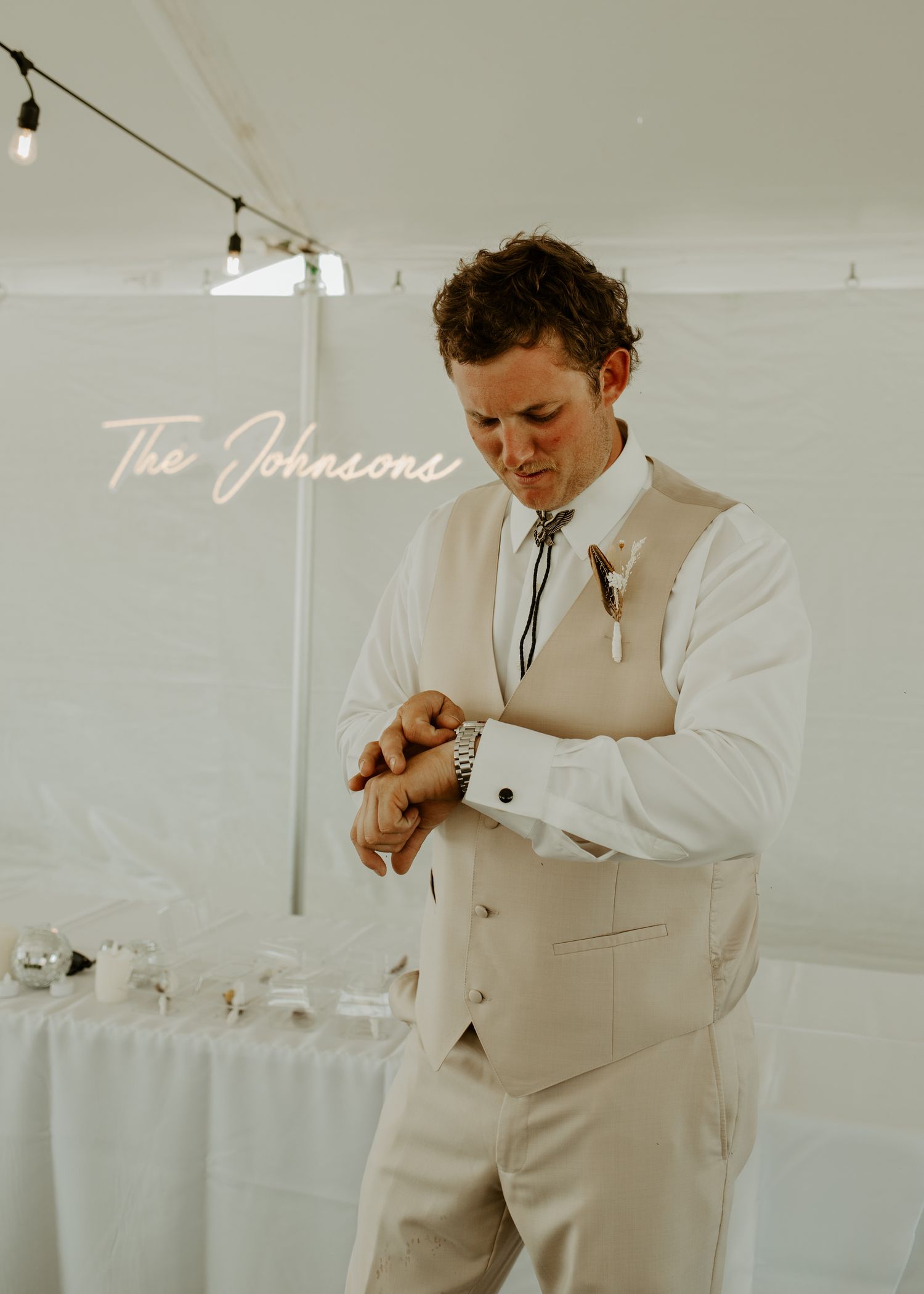 Person in light formal vest checks watch under string lights at wedding venue.