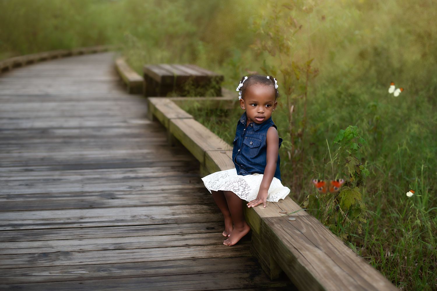 Small barefoot child in dress sitting on wooden boardwalk surrounded by nature.
