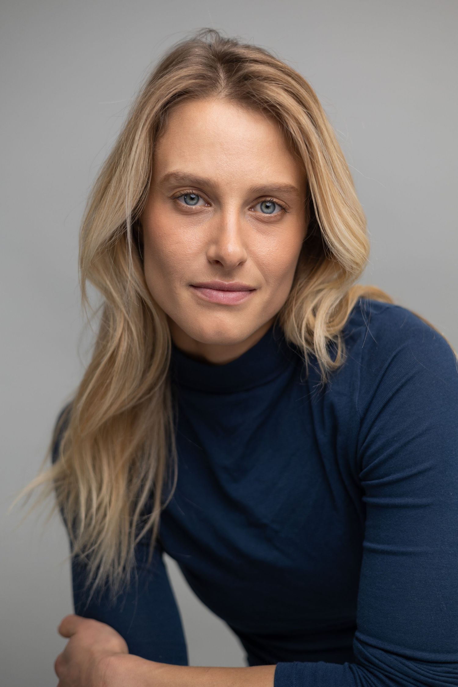 Professional headshot of person in navy turtleneck with blonde hair against grey background.