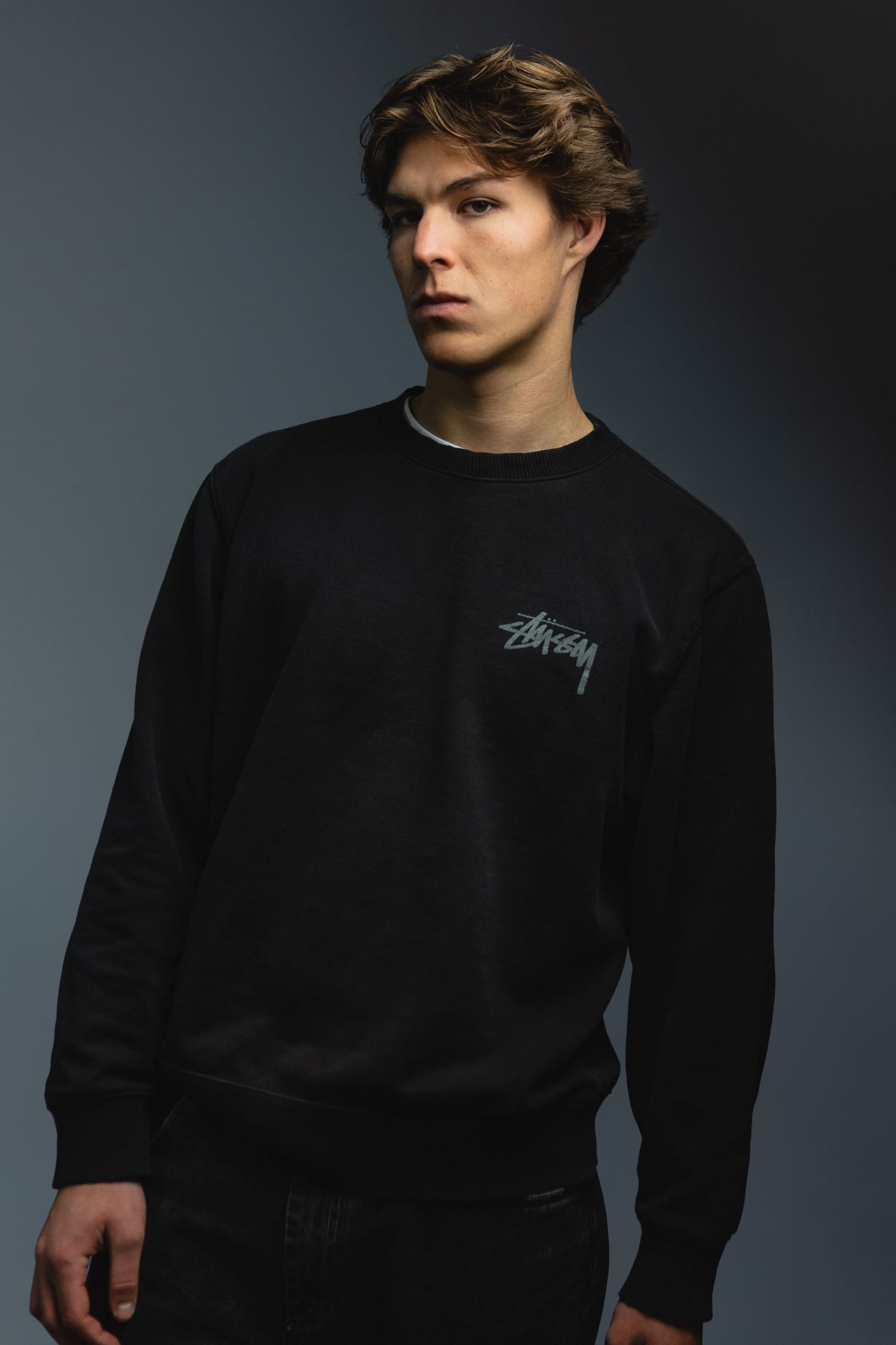 Portrait of person wearing black Stussy sweatshirt against dark gradient background.