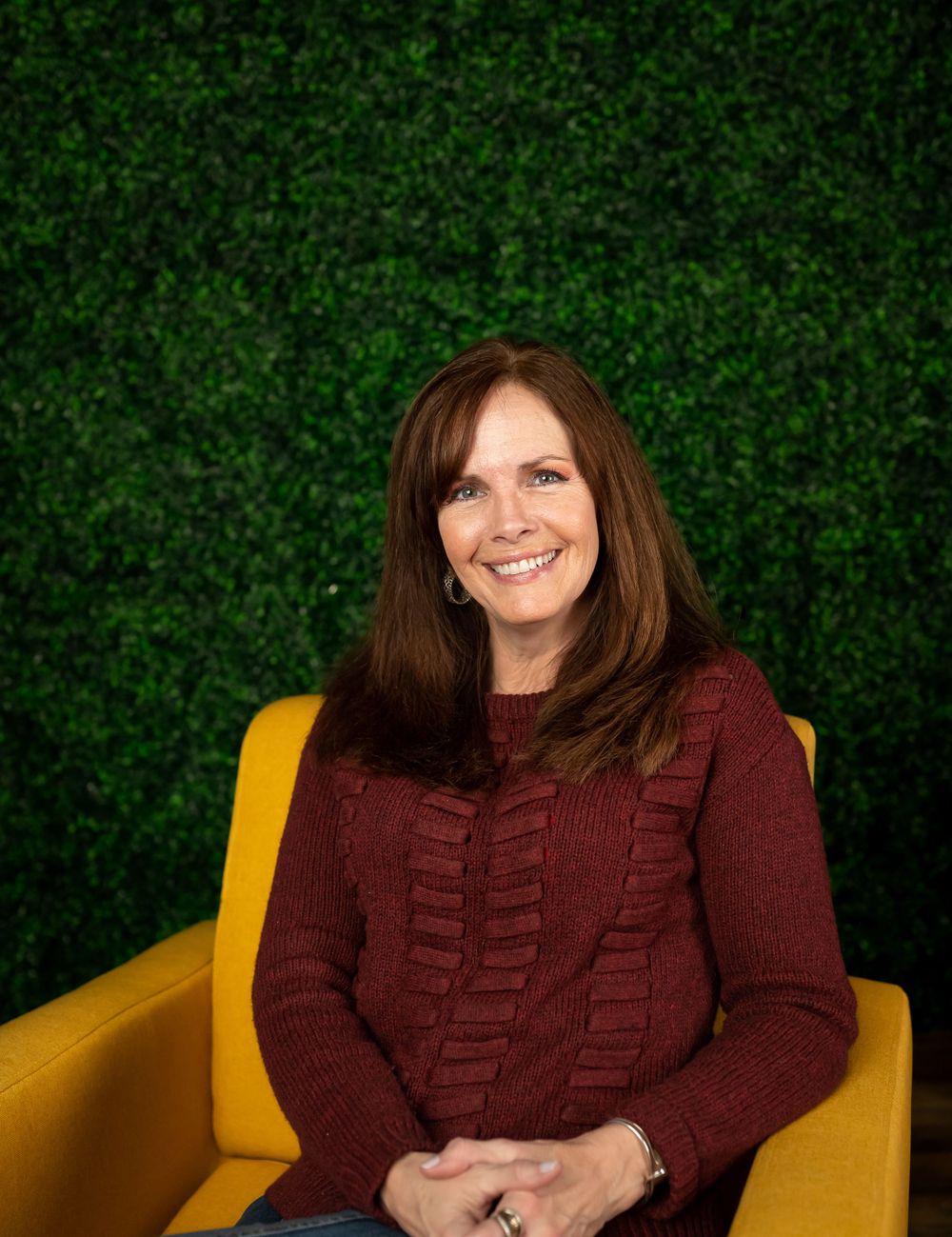 Professional portrait with burgundy sweater seated in yellow chair against green wall.