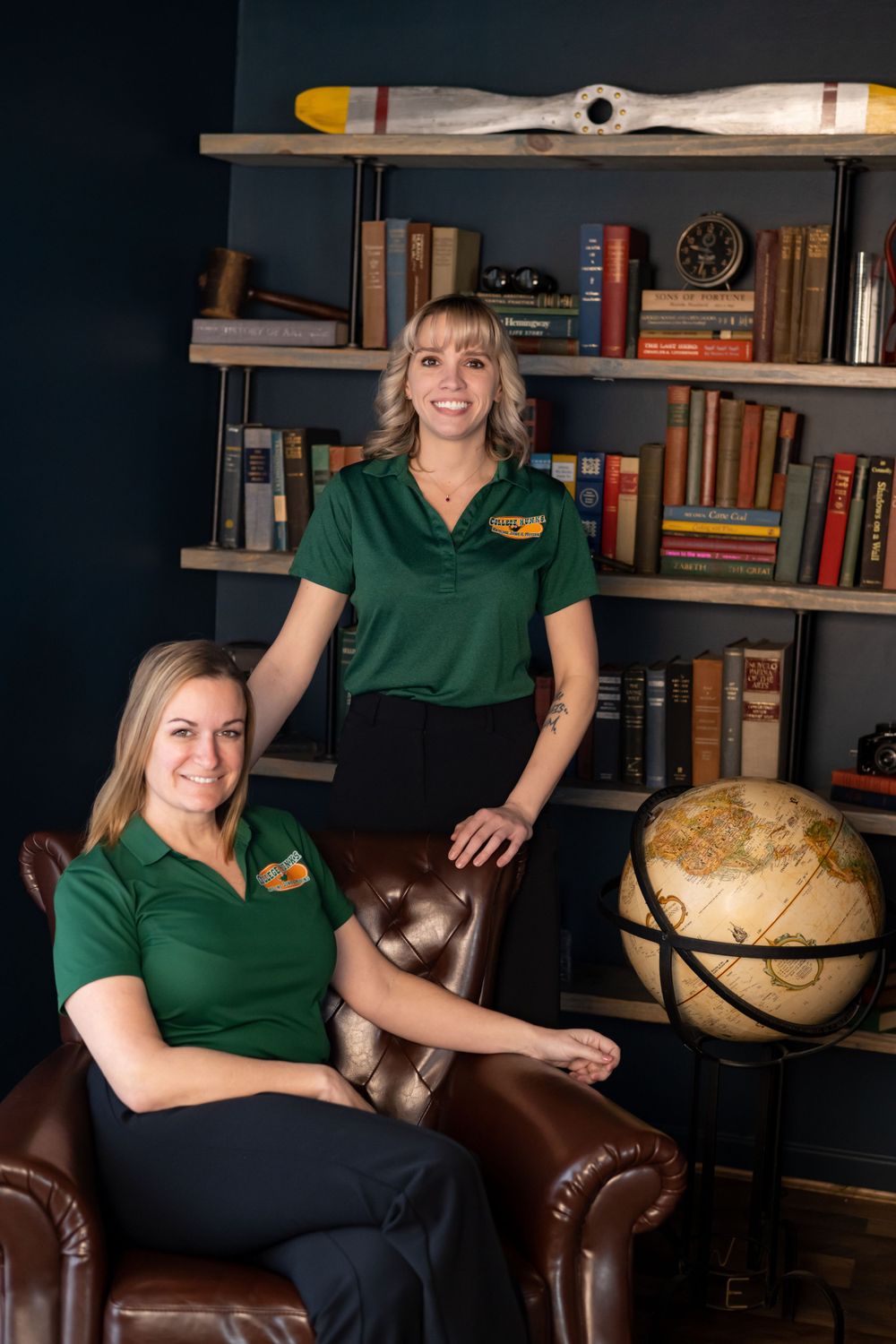 Two staff members in green polo shirts posed in library setting with globe and vintage decor.