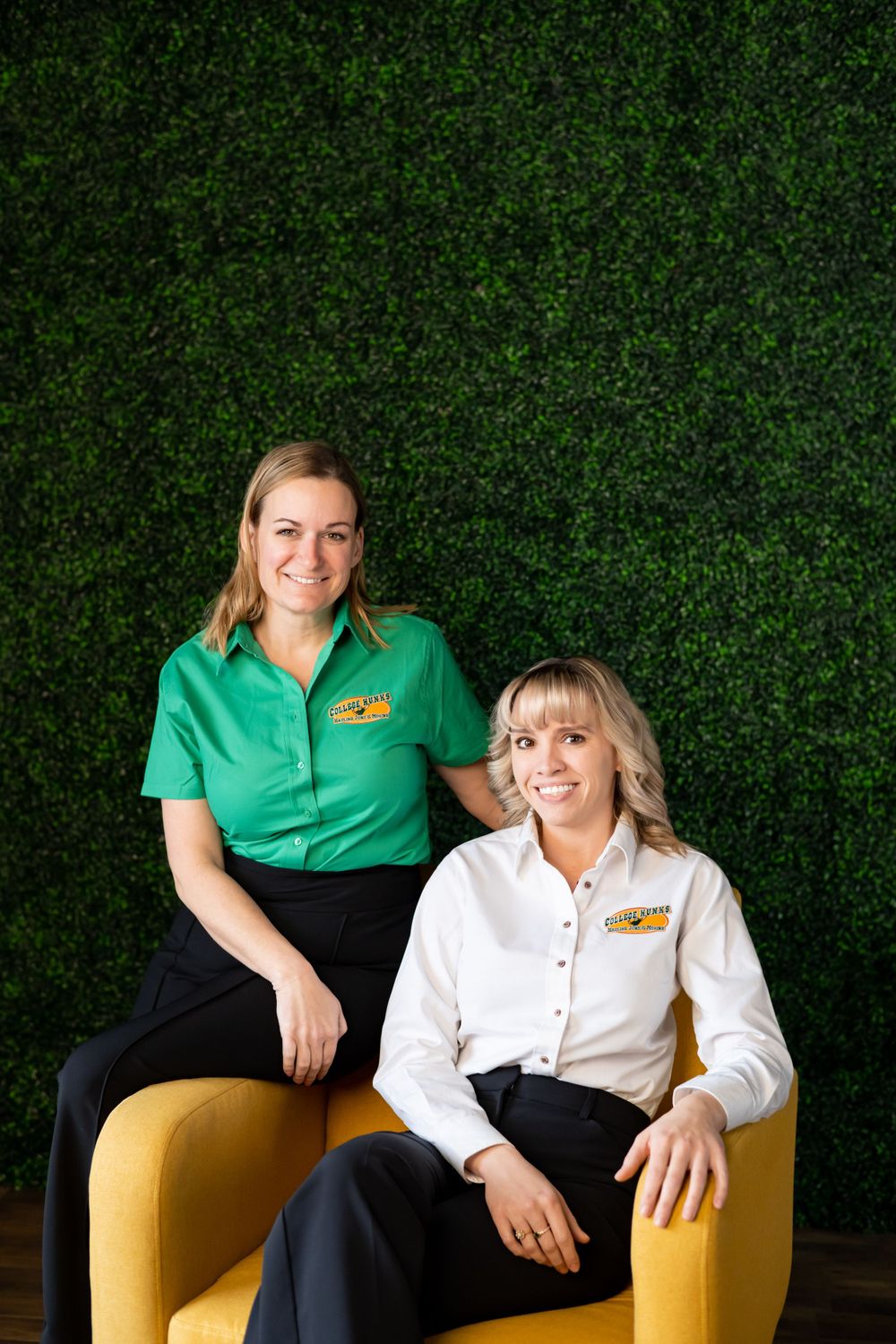 Two staff members in branded uniforms posed on yellow chairs against a green hedge wall.