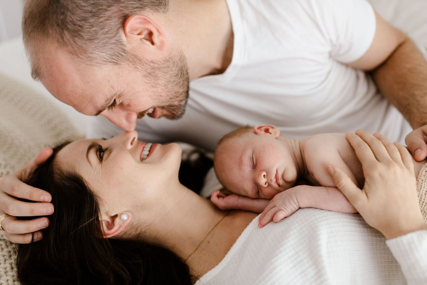 An intimate family moment as parents cuddle with their sleeping newborn on white bedding.