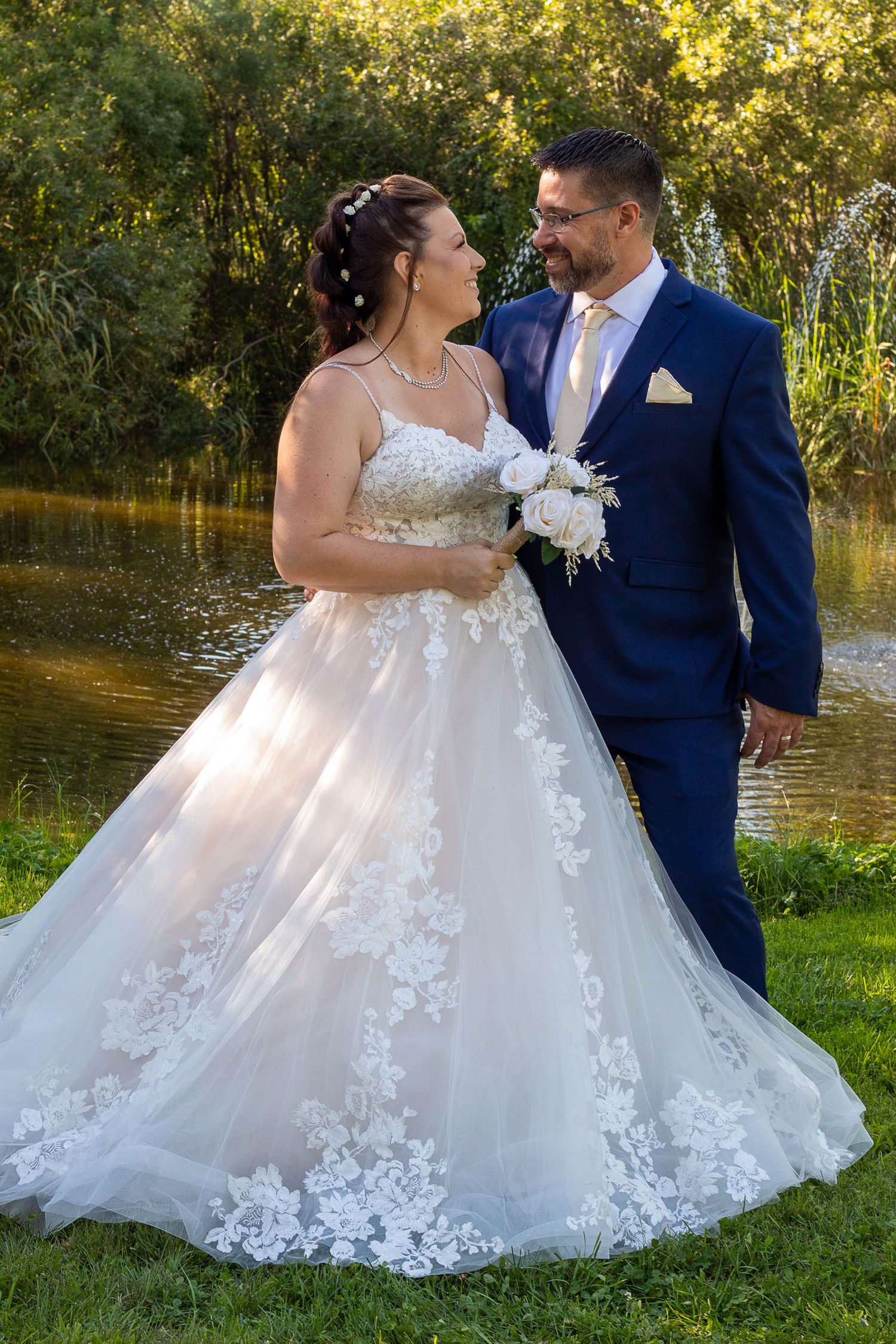 A romantic outdoor wedding portrait by a pond with autumn foliage in the background.