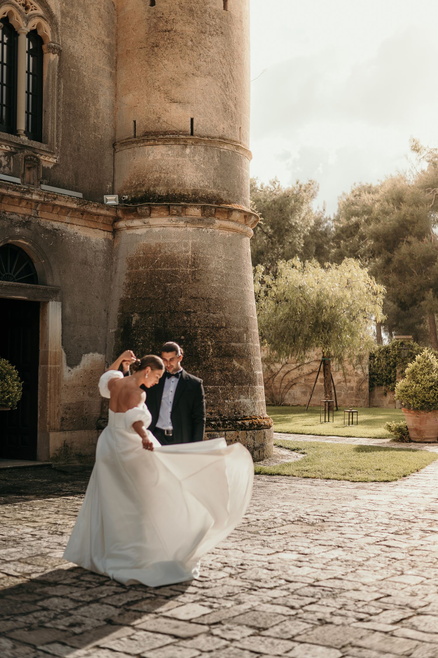 Couple in wedding attire dancing outside historic stone castle with flowing white dress in sunlight.