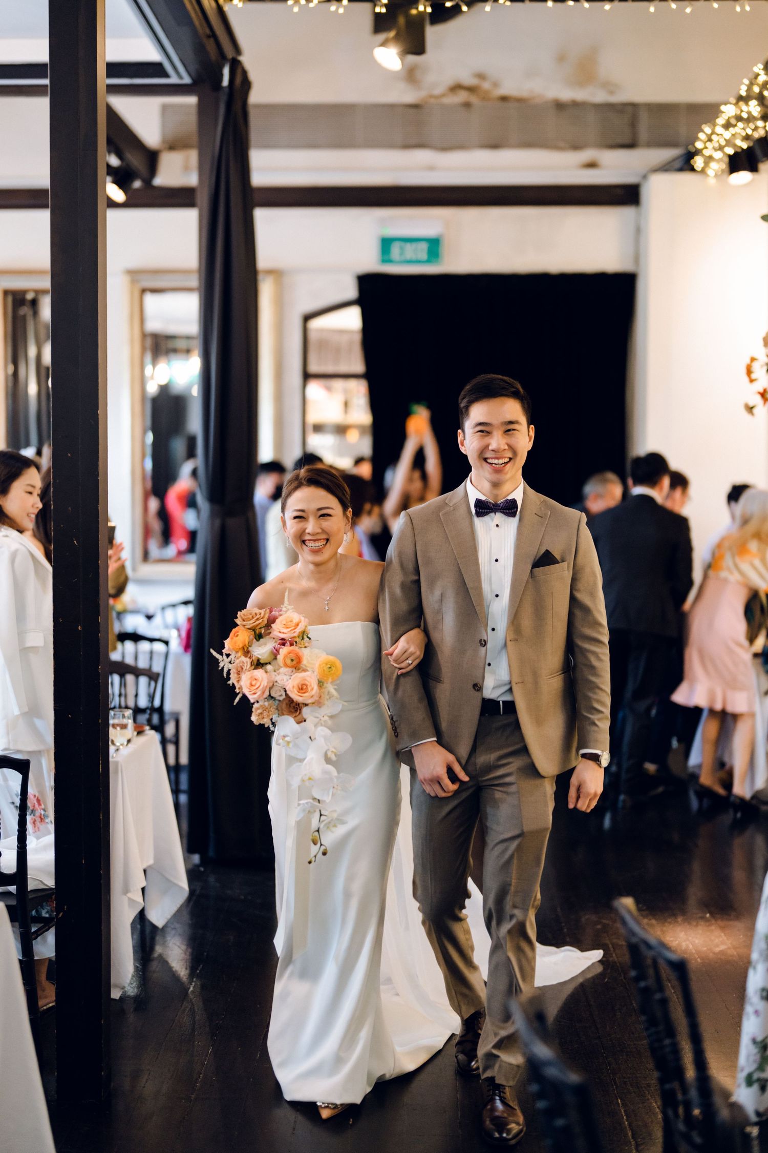 Wedding party members walk joyfully down a bright indoor venue hallway decorated with string lights.