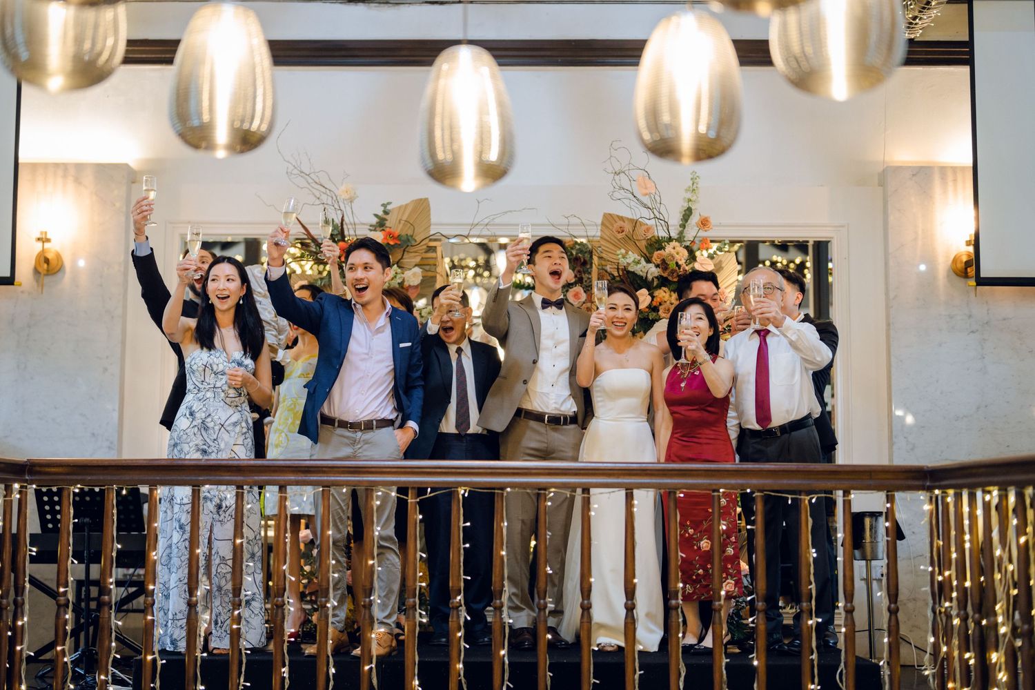 Wedding guests cheer and celebrate during a reception in an elegantly decorated venue with hanging lights.