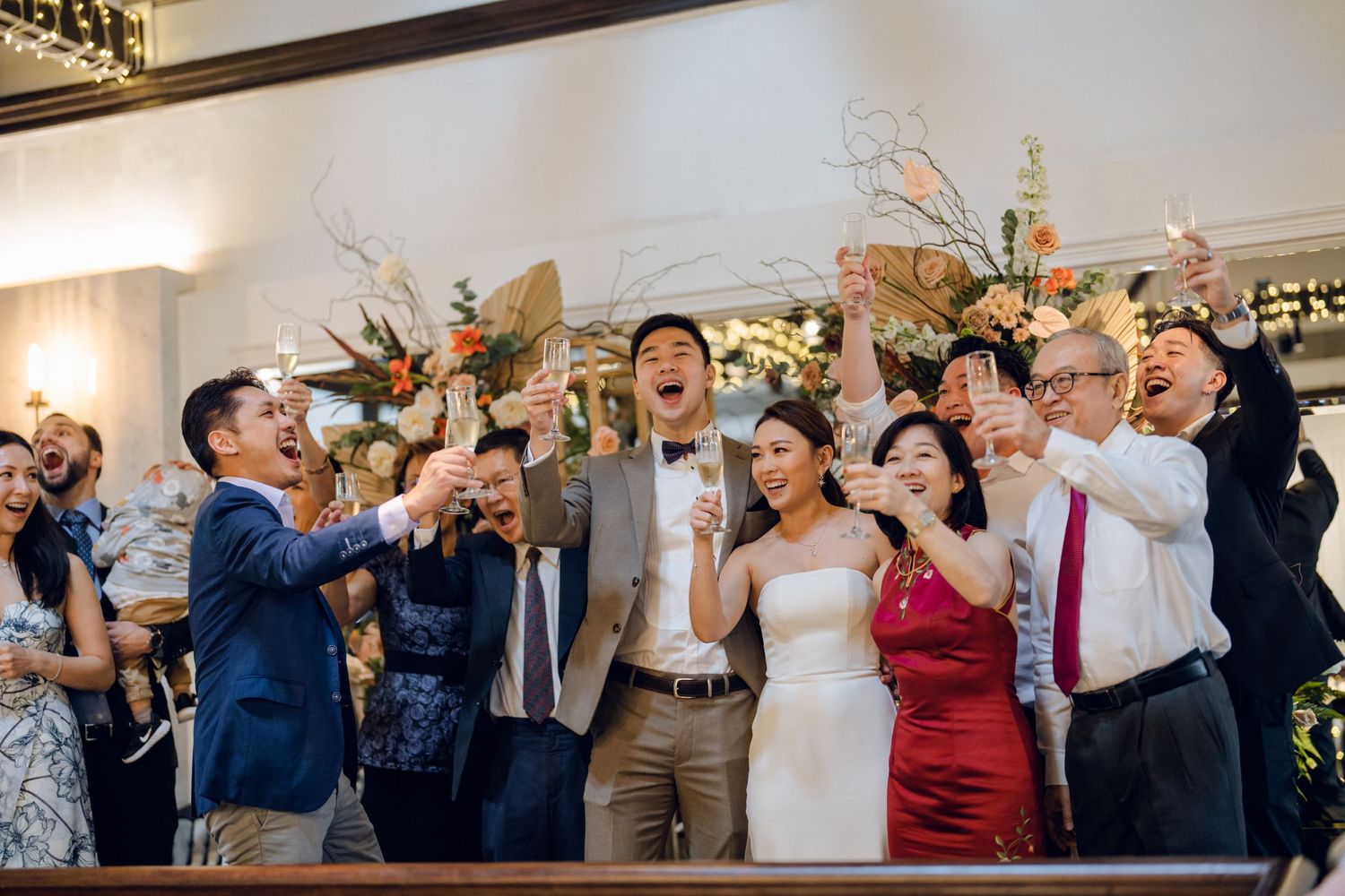 Wedding party and guests celebrating with raised arms in a beautifully decorated reception hall.