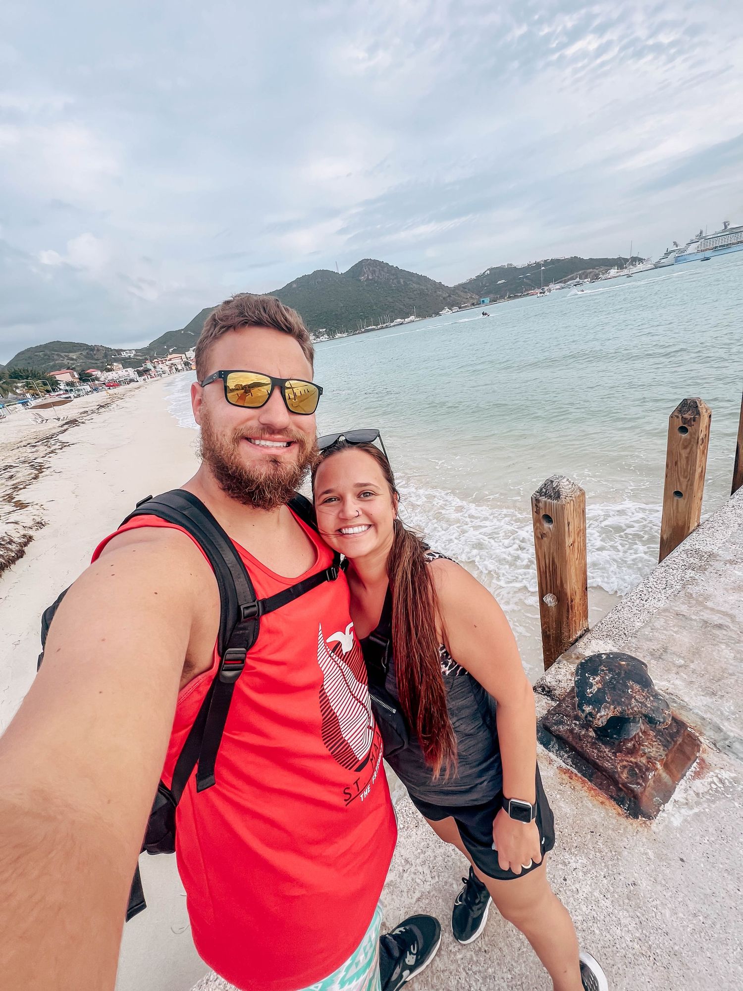 A couple takes a beach selfie with mountains and ocean in the background.