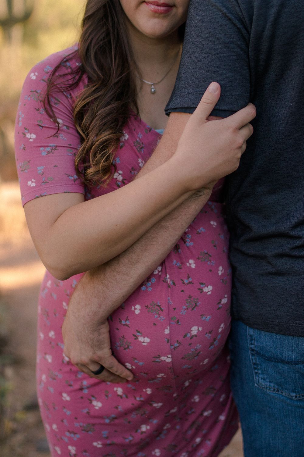 Close-up of expectant couple embracing in pink floral dress against desert scenery.