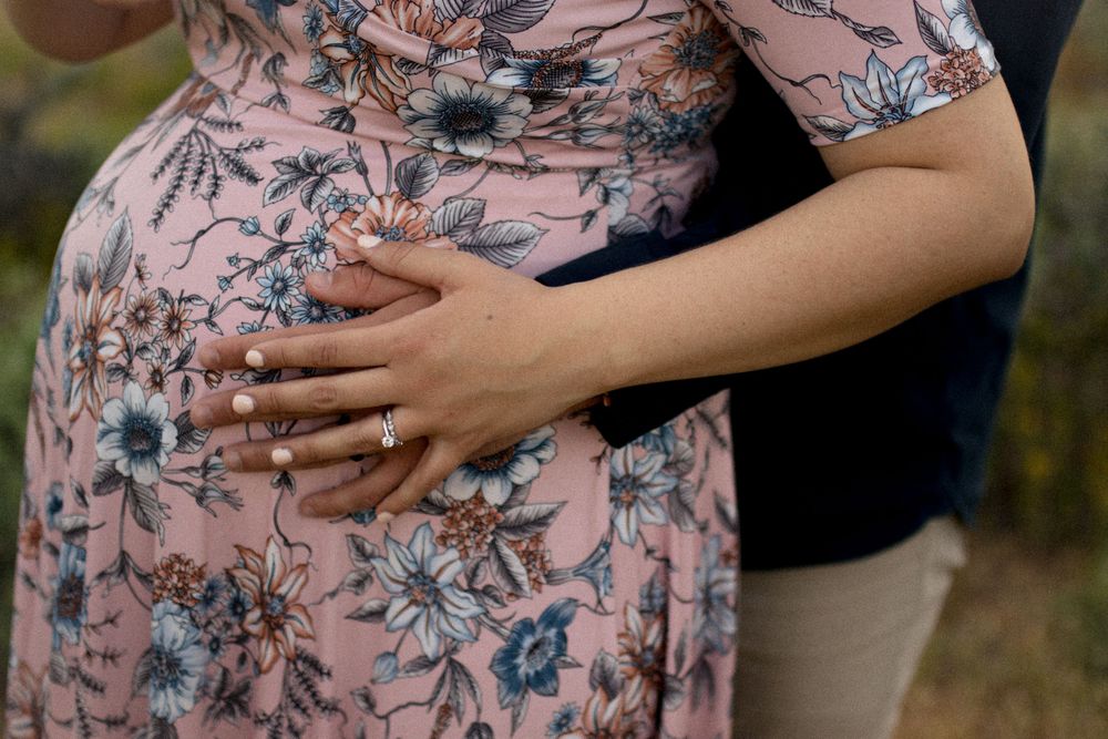Hands rest lovingly on floral maternity dress showing engagement ring.