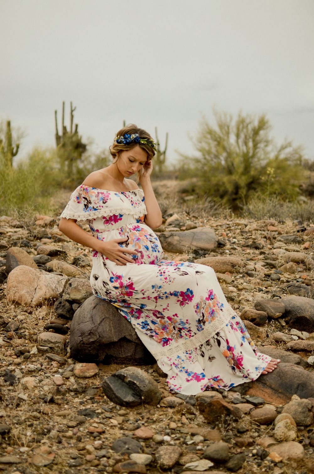A woman in floral dress sits contemplatively on desert rocks surrounded by saguaro cacti.