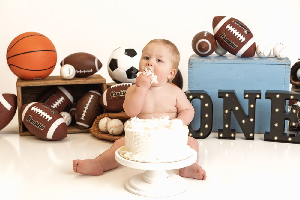 A sports-themed cake smash setup with basketballs and footballs surrounding a white cake on a pedestal.