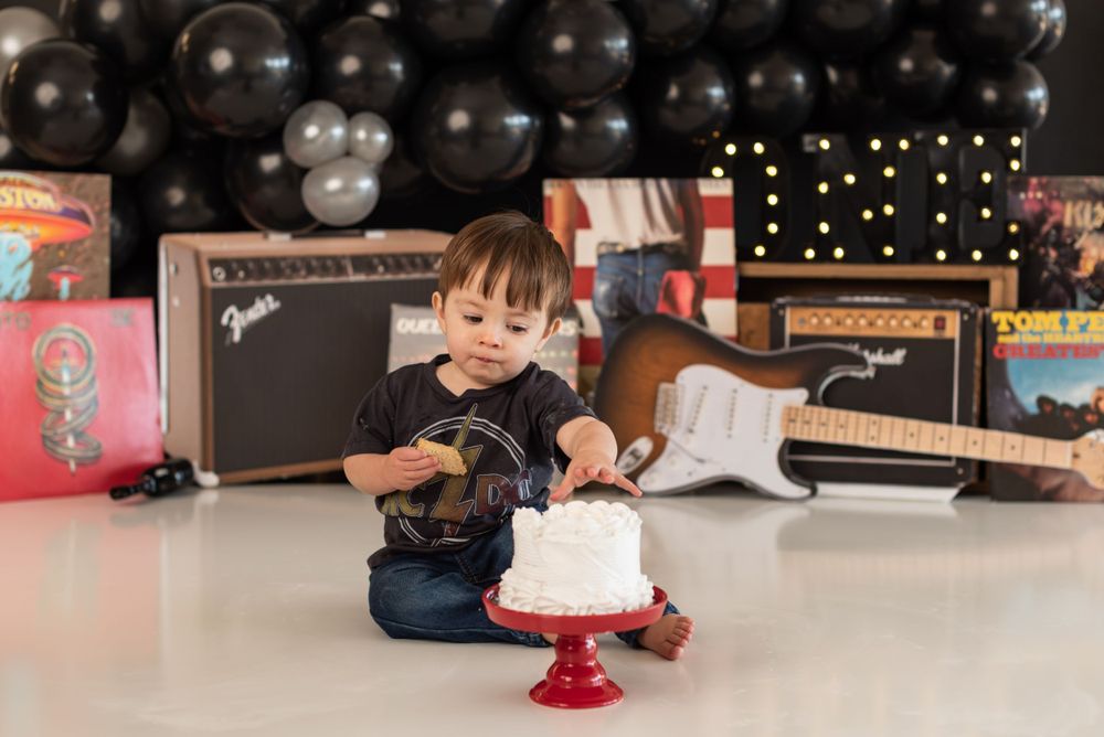 Rock and roll themed birthday cake smash with guitars, amplifiers and black balloons in the background.