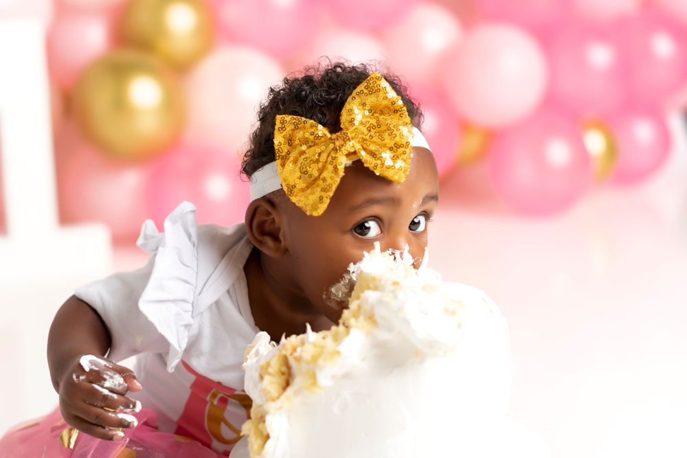 Toddler wearing a gold bow enjoying cake with pink and gold balloon backdrop.
