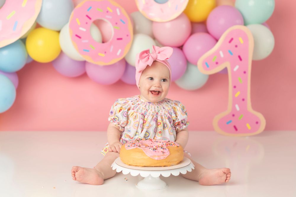 First birthday celebration setup with pink donuts, balloons, and smiling baby with cake.