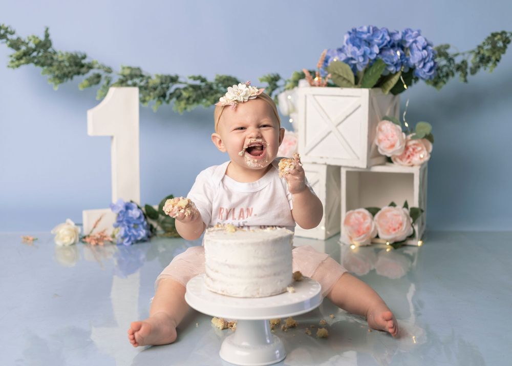 First birthday cake smash with floral decorations and white cake on pedestal against blue background.