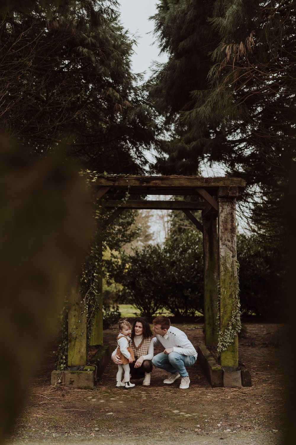 Family crouching together under moss-covered wooden archway in dappled forest light.