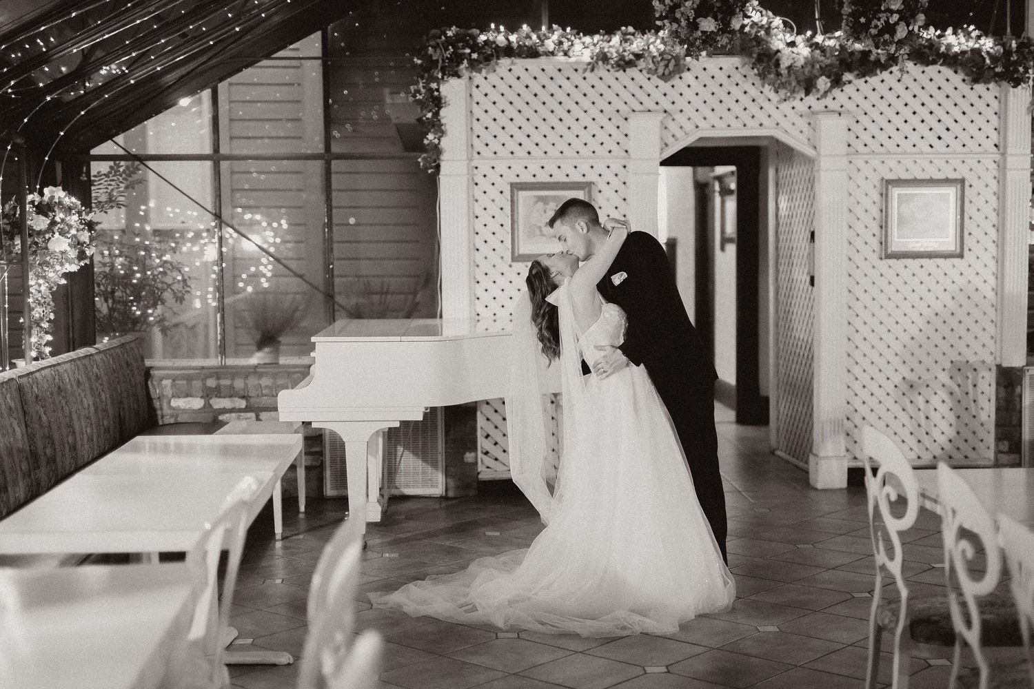 A romantic black and white wedding photo showing a couple dancing near a white piano in a vintage venue.