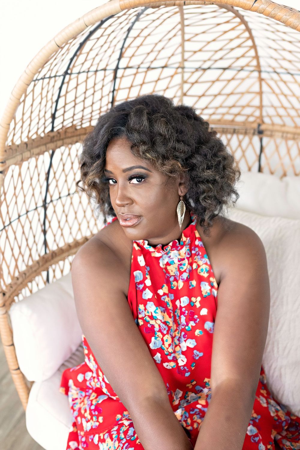 Woman in red floral halter dress sitting in a wicker egg chair with curly hair and gold earrings.