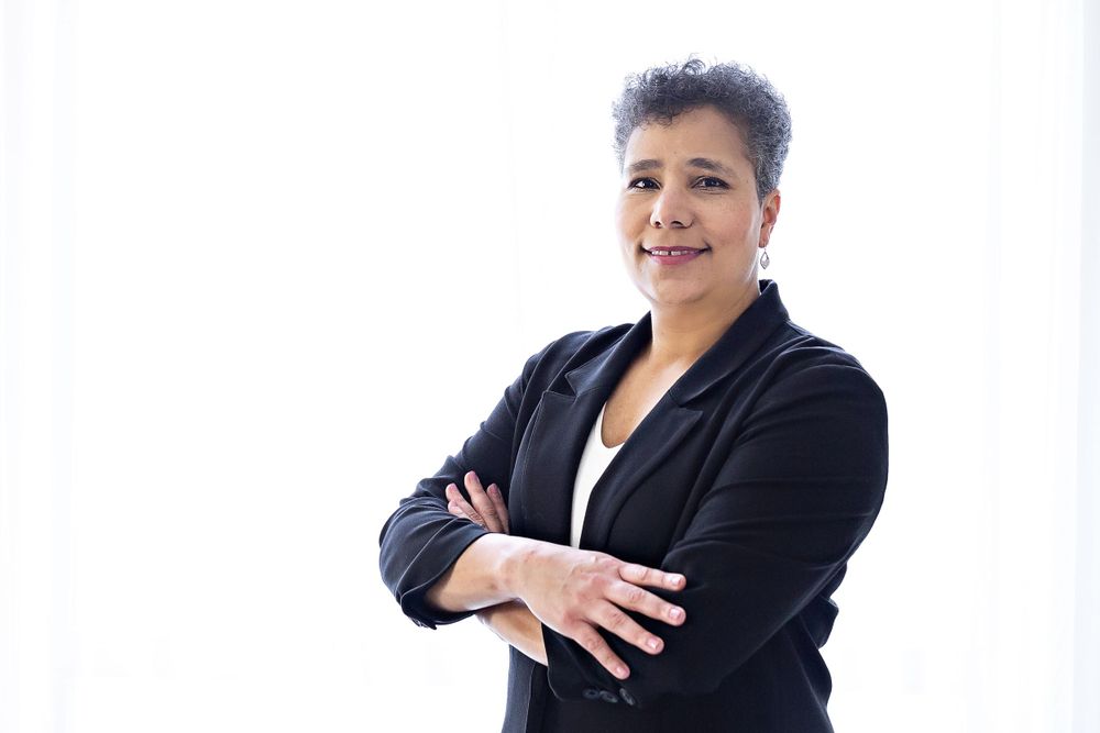 Confident woman in black blazer standing with arms crossed against white background.