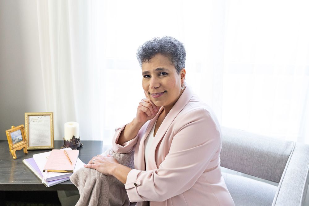 Woman in pink blazer sitting at desk with notebook, posing thoughtfully by window.