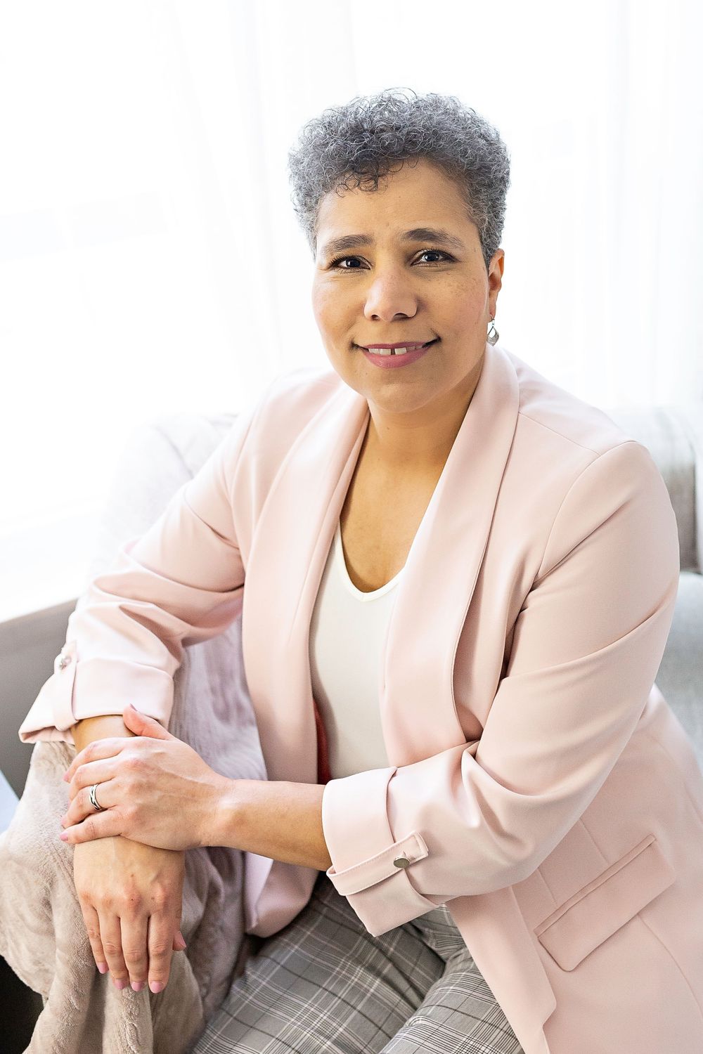 Mature woman in pink blazer sitting relaxed on sofa, smiling warmly at camera.