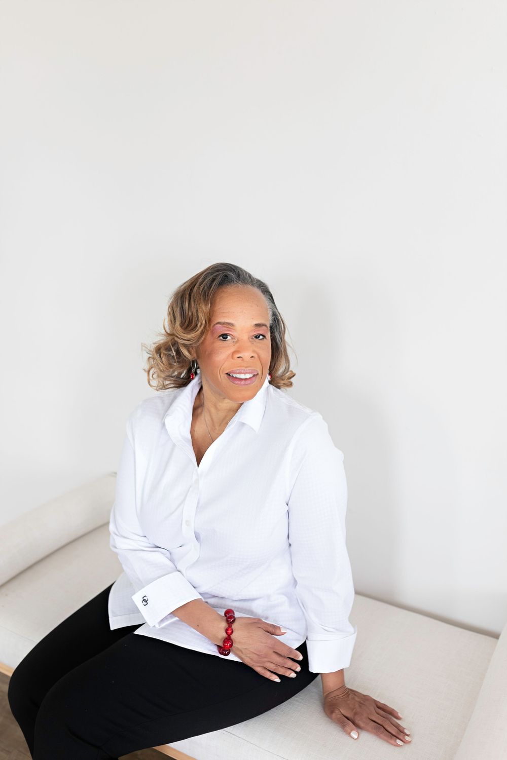 Elegant woman in white button-up shirt sitting on couch with red bracelet and earrings.