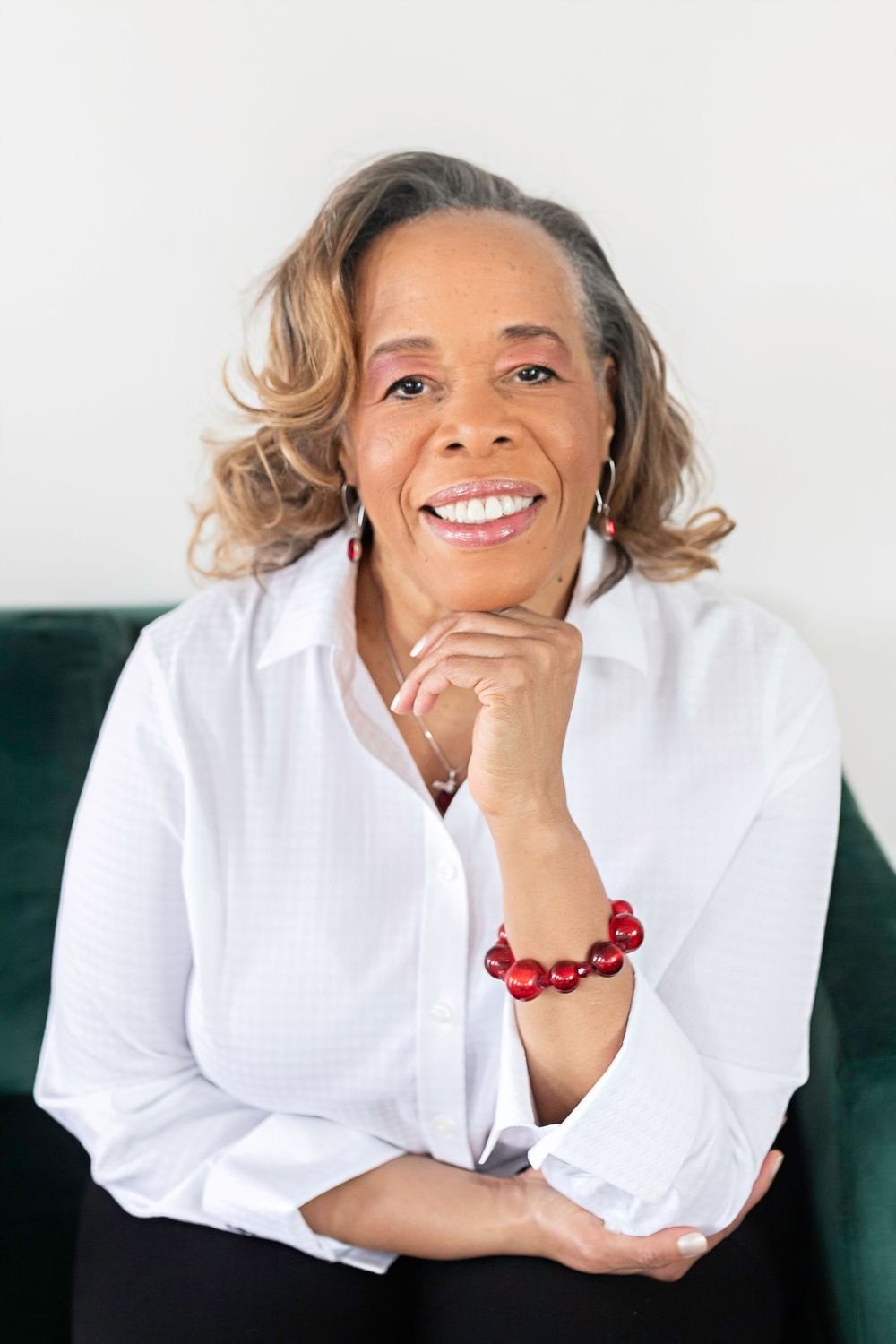 Cheerful woman in white shirt with red bracelet posing on green velvet sofa, smiling.