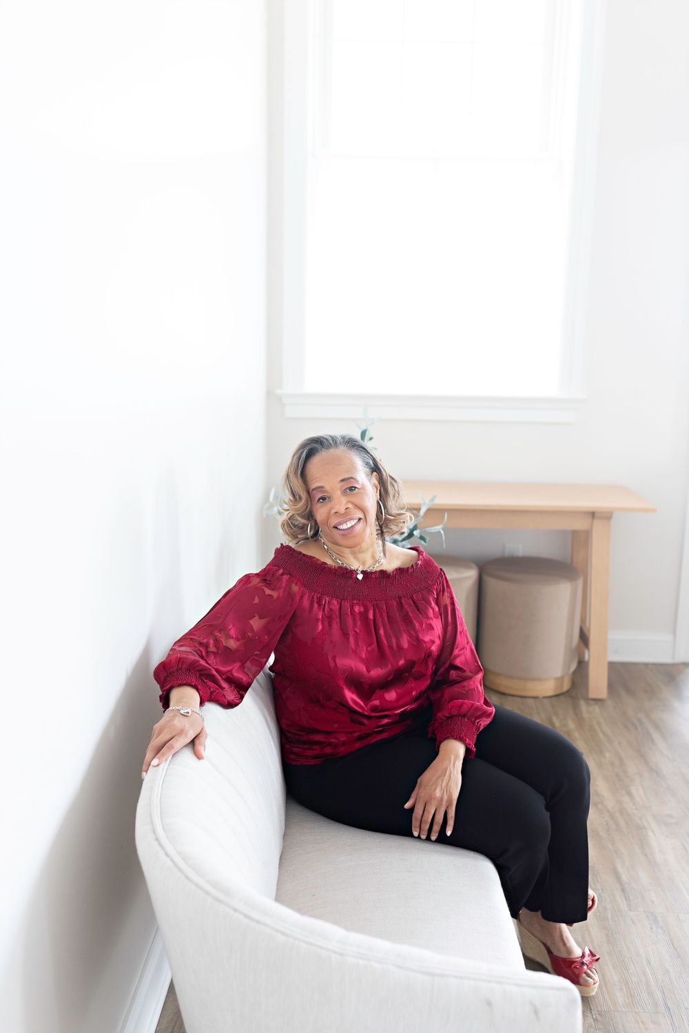 Smiling woman in red off-shoulder blouse sitting in white armchair in bright room.