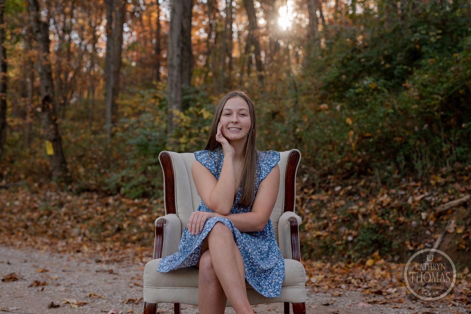 Golden sunlight filters through autumn trees as someone poses in a chair on a leaf-covered path.