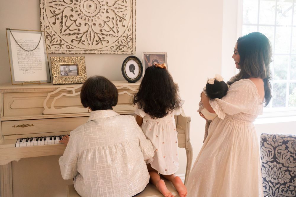 Family gathered at piano wearing white outfits with decorative wall art visible in background.