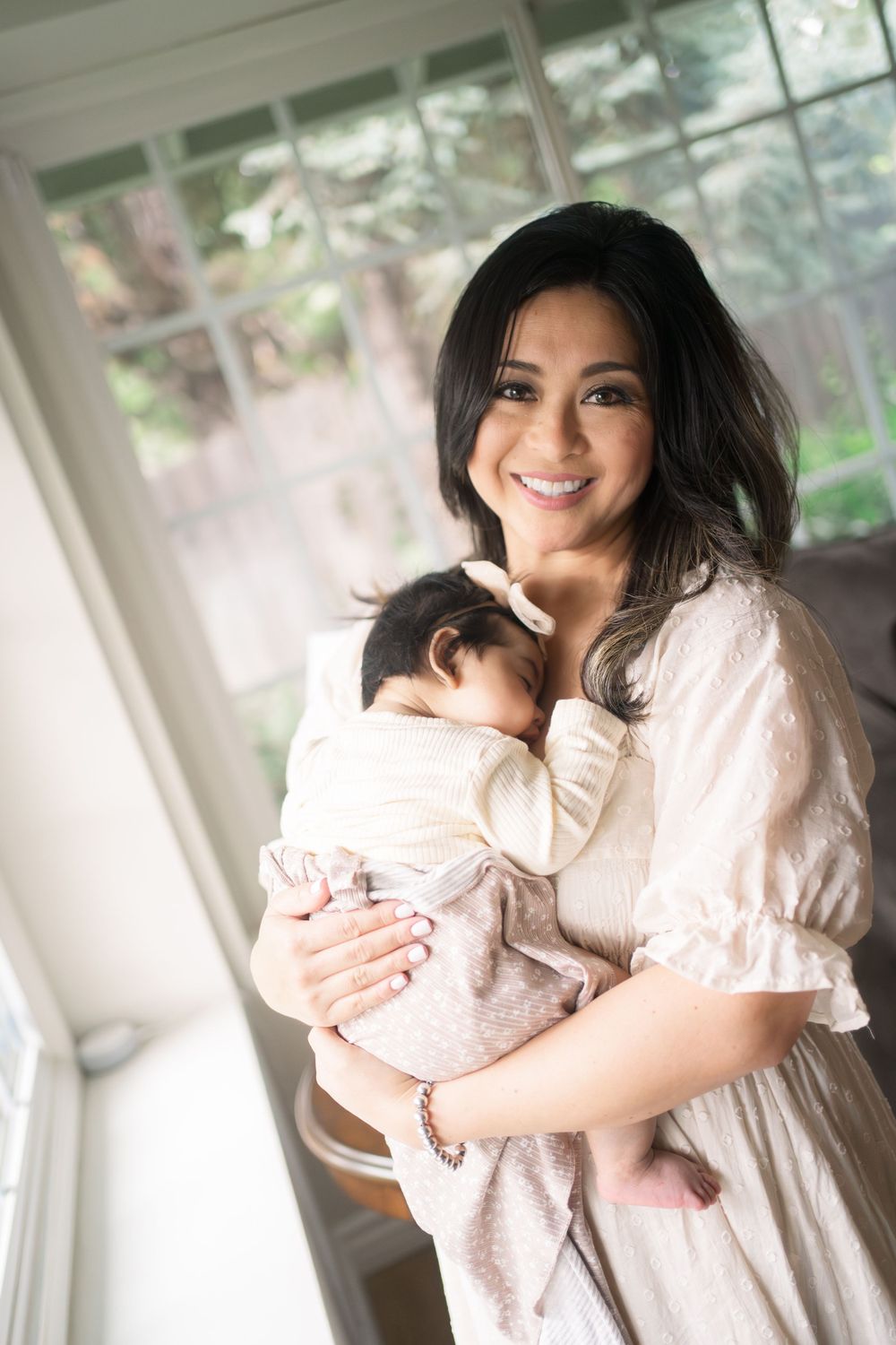A parent in a white dress tenderly holds and smiles at a sleeping baby by a sunlit window.