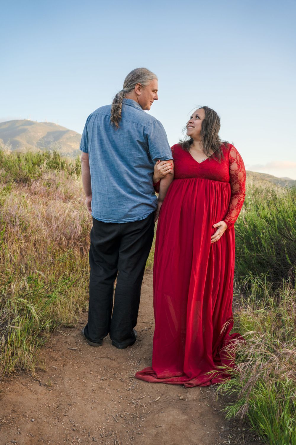 A couple stands together on a dirt trail, with the person in red wearing a flowing dress against mountainous backdrop.