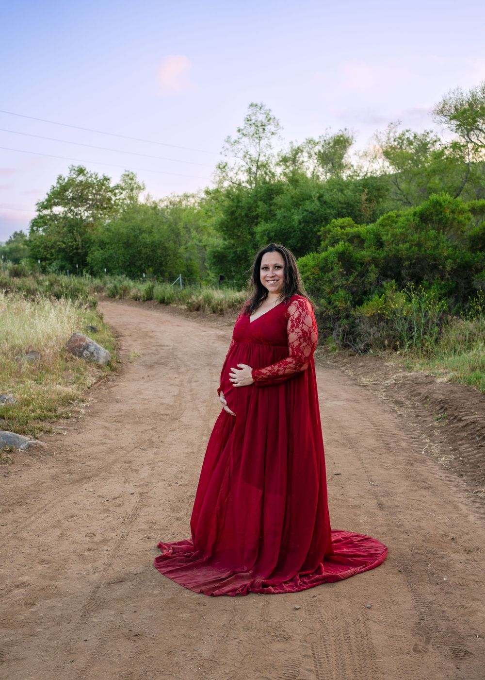 A pregnant figure in a flowing burgundy lace gown poses on a dirt path surrounded by lush greenery at sunset.