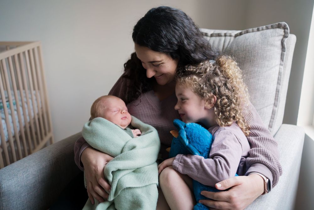 A tender family moment on a couch as someone holds a swaddled baby while a curly-haired child looks on.