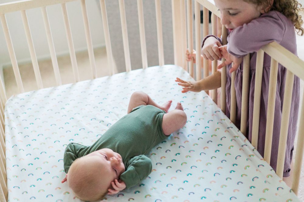 A baby in green sleepwear lies on a crib mattress with a patterned sheet while a person looks from above.