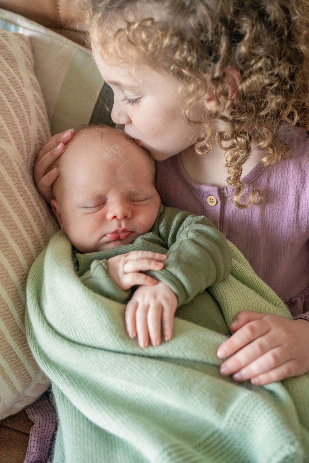 A toddler with curly hair gives a gentle kiss to a sleeping newborn baby wrapped in mint green blankets.