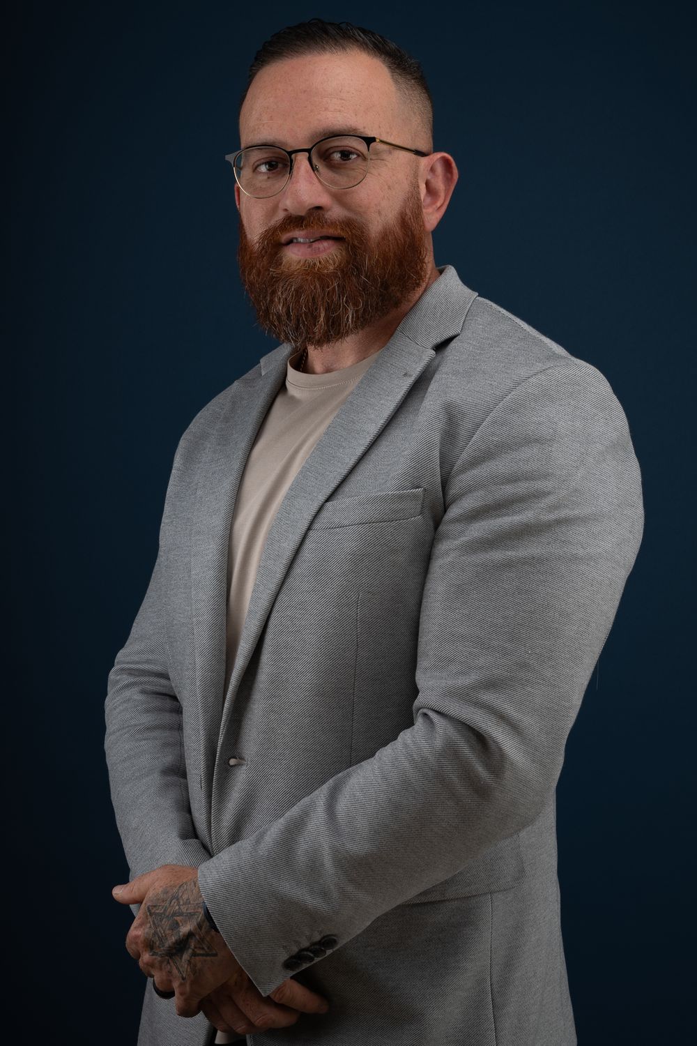 Professional headshot of person wearing glasses and grey blazer against dark background.