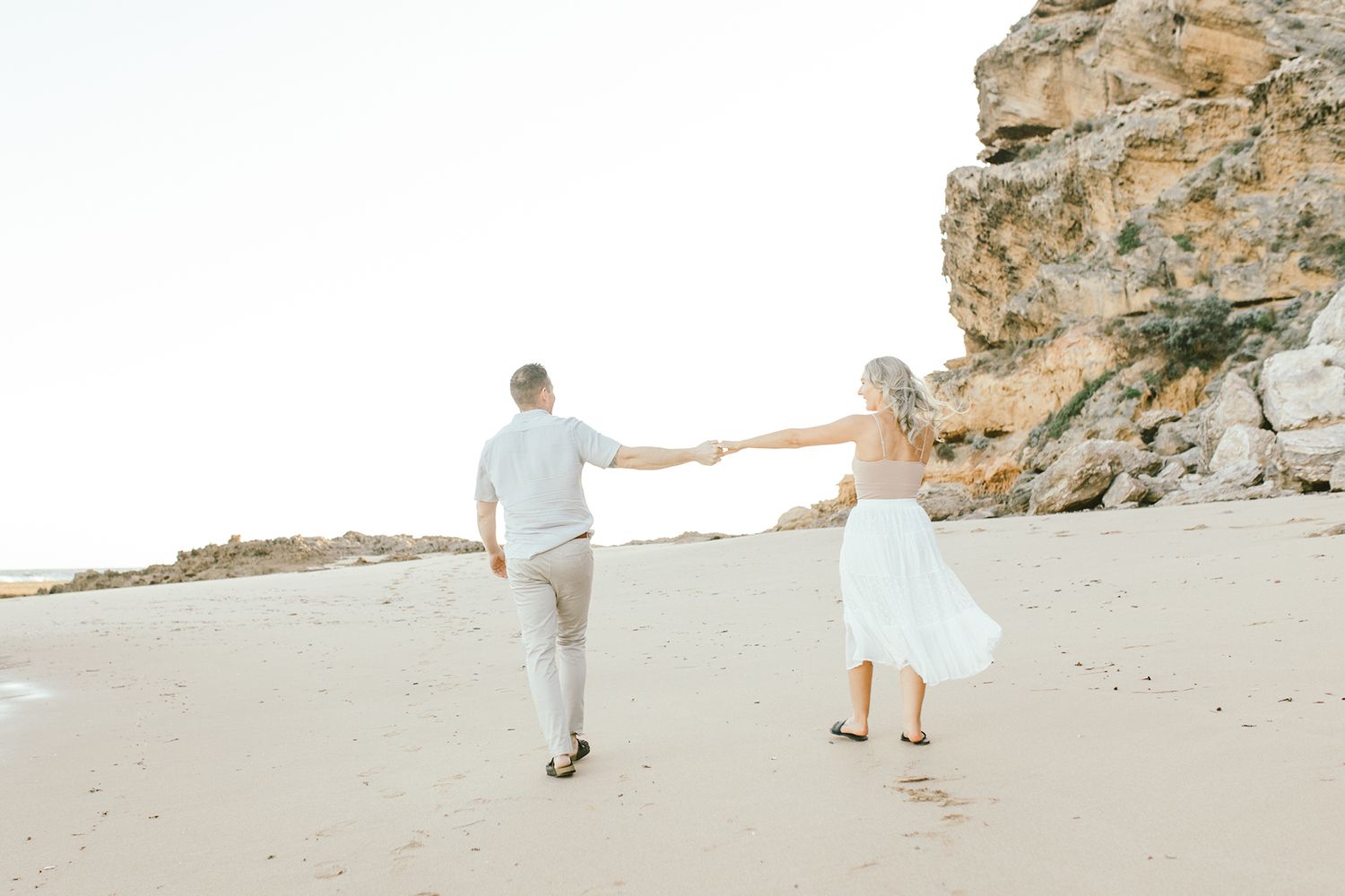 Couple holding hands while walking on a secluded beach with rocky cliffs.