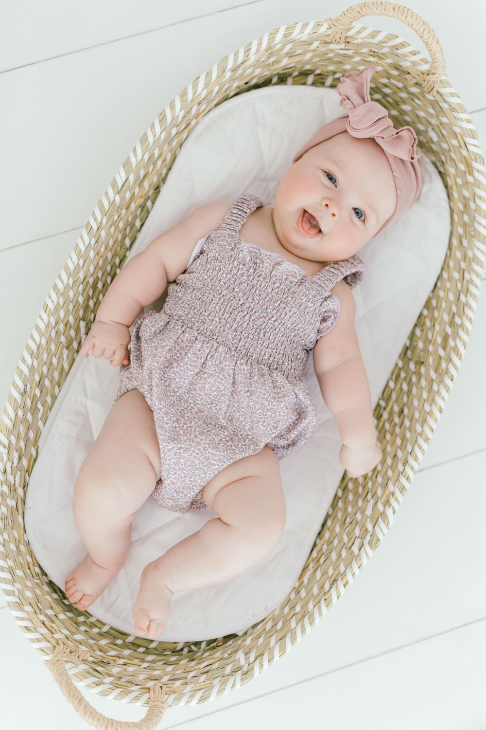 Smiling baby in pink romper and headband resting in woven basket on white background.