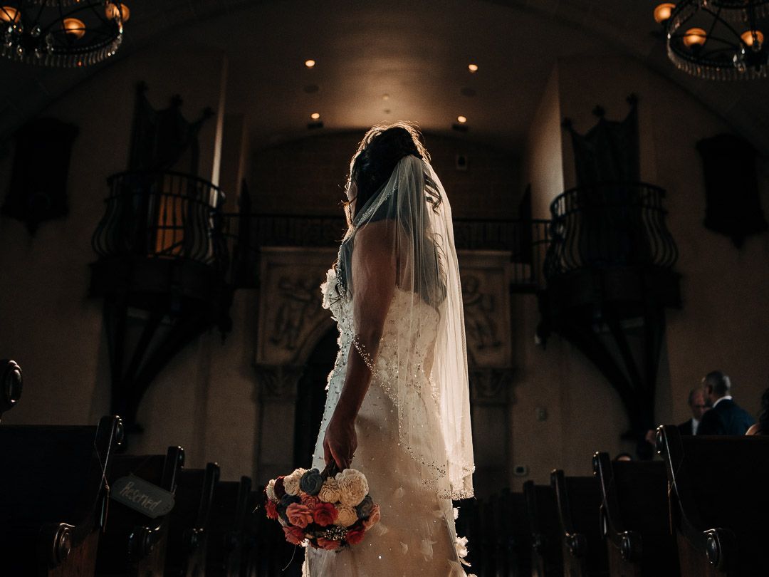 A dramatic silhouette of a bride holding flowers in a church with glowing chandeliers.