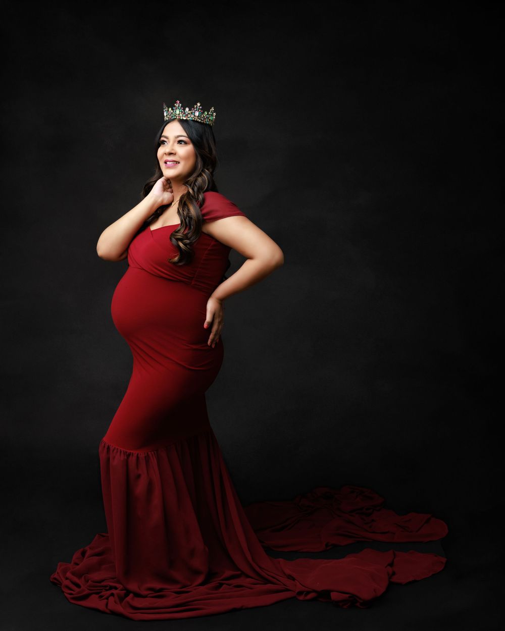 Maternity portrait in flowing red gown and crown against dark background with dramatic lighting.