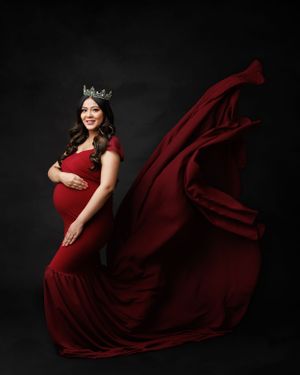 A dramatic maternity portrait in a flowing red dress with crown against black background.